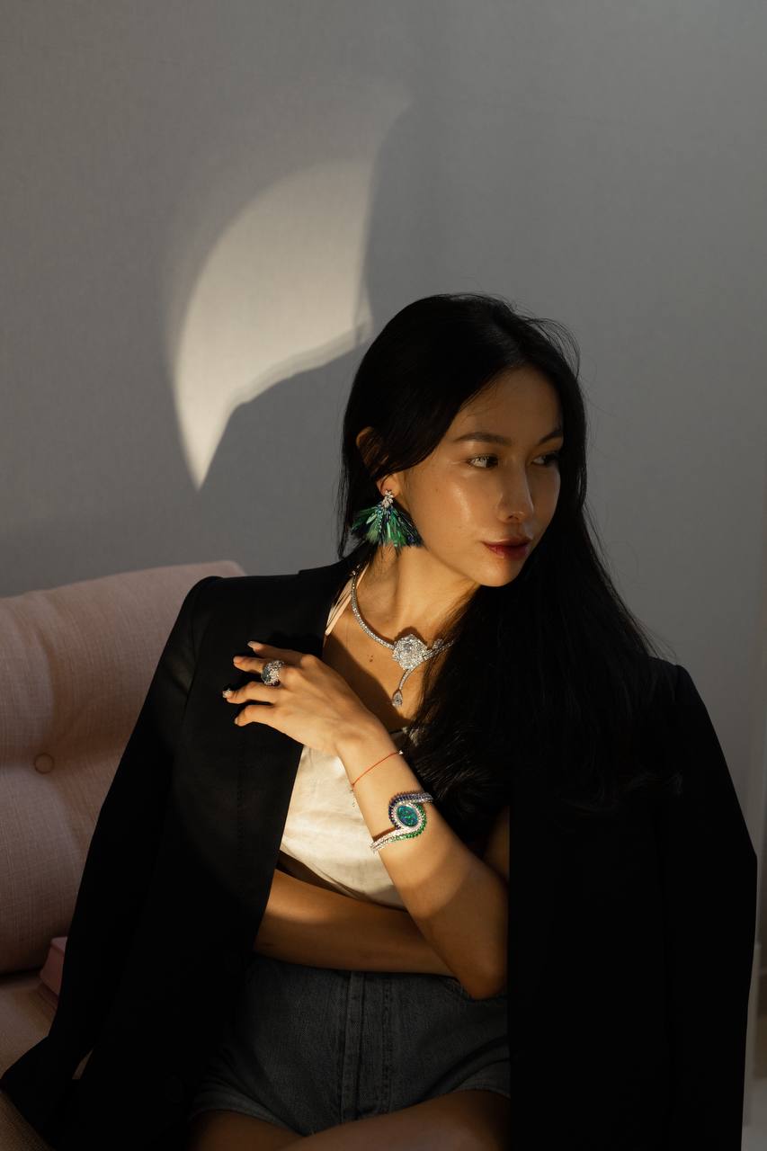 Chelsea seated in a black jacket, looking to the side with one hand near her necklace and a green earring